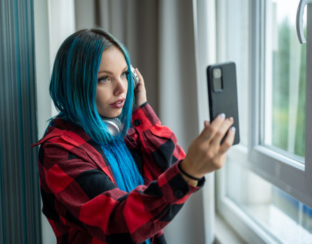 Portrait of beautiful blue haired caucasian hipster woman standing in front of window taking selfie with her mobile phone. Wearing headphones listening to music. Using technology.の写真素材