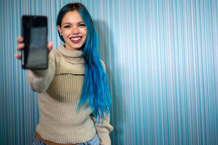 Portrait of a beautiful blue-haired hipster woman receiving a notification that causes her to have an extremely positive and surprised reaction. She extends her arm with her mobile phone towards us.の写真素材
