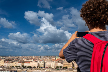 Budapest,Hungary,August 29,2022. A young man takes pictures with his mobile phone of the magnificent cityscape from the top of the hill. Beautiful summer day with blue sky and white clouds.の写真素材