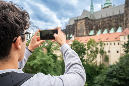 Prague, Czech Republic, August 8, 2023. A young man takes photos with his cell phone of the view of Prague Castle and St. Vitus Cathedral on the hill. A beautiful summer day. Travel destinations.の写真素材
