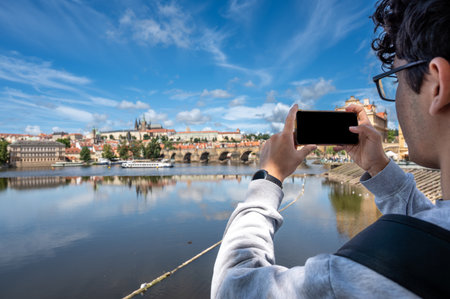 Prague, Czech Republic, August 9, 2023. A young Caucasian man takes photos with his cell phone of the enchanting view of the castle and cathedral dominating the hill. A lifestyle using technology.の写真素材