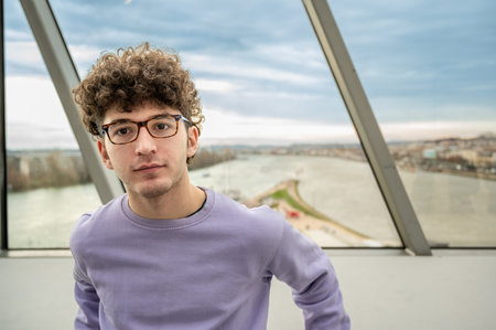 Young man with curly hair and glasses wearing a purple sweater, looking directly at the camera with...