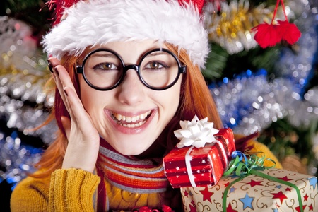 Funny girl in christmas cap with gift boxes and tree at background.