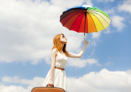Beautiful redhead girl with umbrella and suitcase at blue sky background.の写真素材