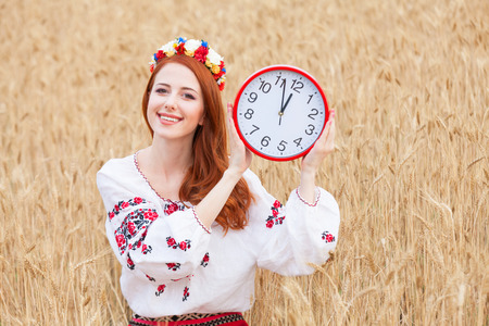 Redhead girl in national ukrainian clothes with clock on the wheat field.