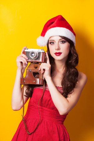 Beautiful brunette girl in christmas hat with camera on yellow background.の写真素材