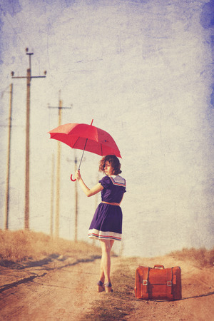 Beautiful brunette girl with suitcase and umbrella on the countryside road.の写真素材