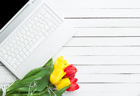 White computer and bouquet of tulips on a white wooden table.の写真素材