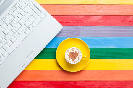Cup of cappuccino with heart shape and computer on multicolor wooden background.の写真素材