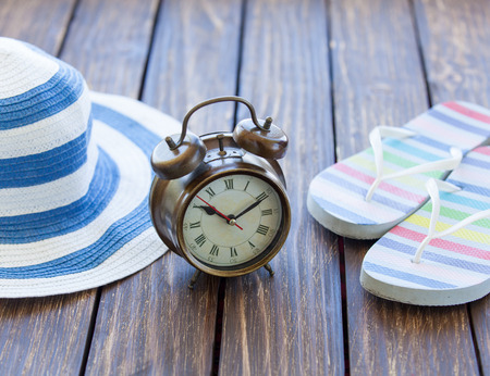 Alarm clock and hat with flip flops on wooden table.