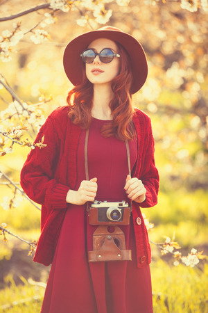 Portrait of a beautiful redhead women in red sweater and hat with camera in blossom apple tree garden in spring time on sunset.の写真素材