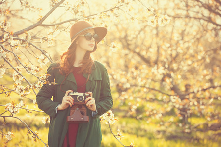 Portrait of a beautiful redhead women in cloak and hat with camera in blossom apple tree garden in spring time on sunset.の写真素材