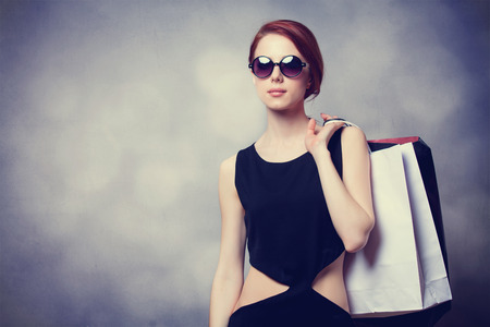 Style redhead girl with sunglasses and black dress with shopping bags on grey backgroundの写真素材