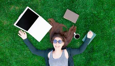 portrait of a beautiful brunette student girl with laptop computer and books on the green grass in...