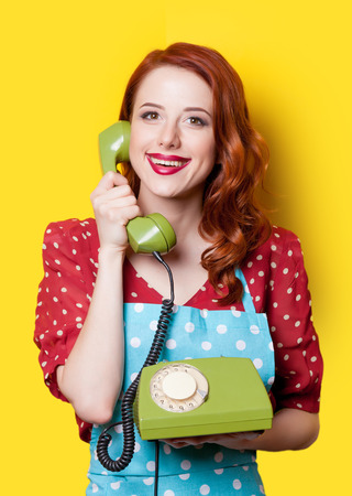 Smiling redhead girl in red polka dot dress and blue apron with green dial phone on yellow background.の写真素材