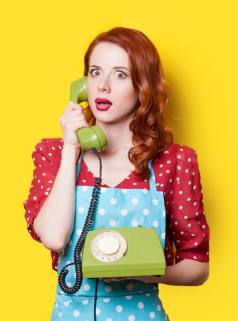 Surprised redhead girl in red polka dot dress and blue apron with green dial phone on yellow background.の写真素材