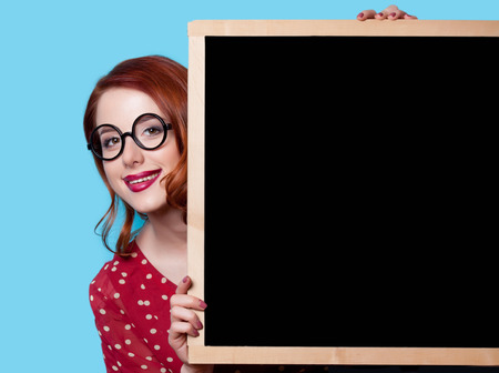 Young smiling redhead teacher in red polka dot dress with blackboard on blue background.の写真素材