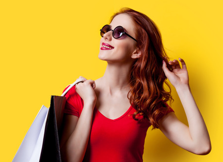 Redhead girl in red dress and sunglasses with shopping bags on yellow backgroundの写真素材