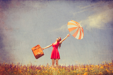 Portrait of a beautiful redhead girl in red dress with umbrella and suitcase on the meadow.の写真素材