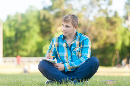 Young teen boy with notebook on the green grass in the park.