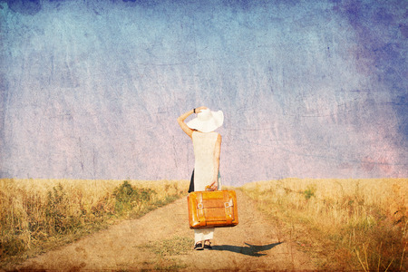 Redhead girl with suitcase at countryside road near wheat field. Photo in old color image style.の写真素材