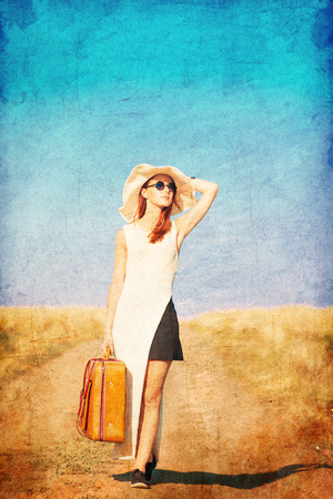 Redhead girl with suitcase at countryside road near wheat field. Photo in old color image style.の写真素材
