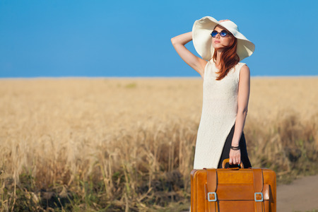 Redhead girl with suitcase at countryside road near wheat field.の写真素材