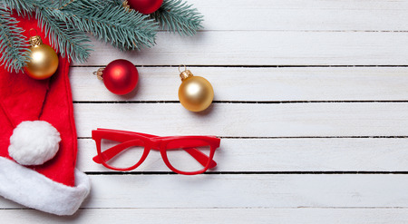 Santas hat and christmas bubbles with red glasses on white wooden table.の写真素材