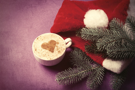 Cup of coffee and fir-tree branch with Santas hat on violet backgorund.の写真素材