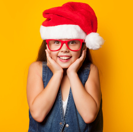Beautiful young girl in red glasses and Santas hat on yellow background.の写真素材