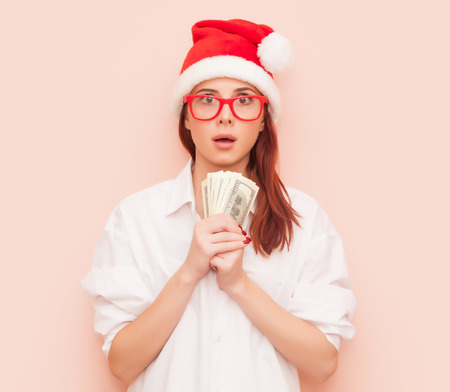 Portrait of surprised redhead woman in Santa Claus hat with money on pink backgroundの写真素材