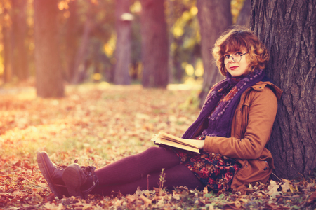 Portrait of a young redhead student girl with book  near tree in the autumn parkの写真素材