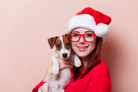 Portrait of a young redhead woman in Santa Claus hat with jack russell terrier puppy on pink backgroundの写真素材