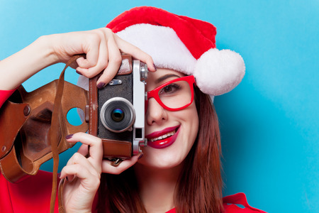 Portrait of a young redhead woman in Santa Claus hat and camera on blue backgroundの写真素材
