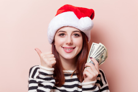 Portrait of a young redhead woman in Santa Claus hat with money on pink backgroundの写真素材