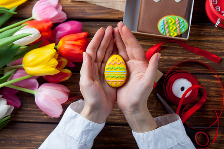 Female hands are holding a Easter cookie egg near flowers before wrapping on violet background