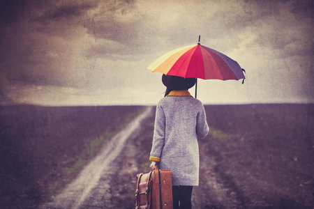 portrait of a young beautiful woman with colorful umbrella and brown suitcase standing on the roadの写真素材