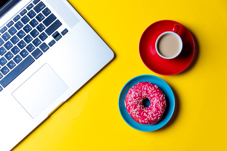 photo of the glazed donut on the plate, cup of coffee and laptop on the yellow backgroundの写真素材
