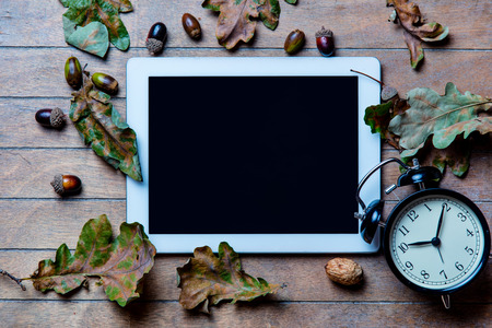 photo of the cool tablet, alarm clock and fallen leaves on the brown wooden background