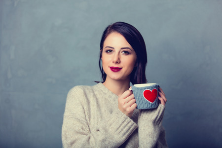 Portrait of a style brunette woman with cup of coffee or tea with heart shape symbol on grey backgroundの写真素材