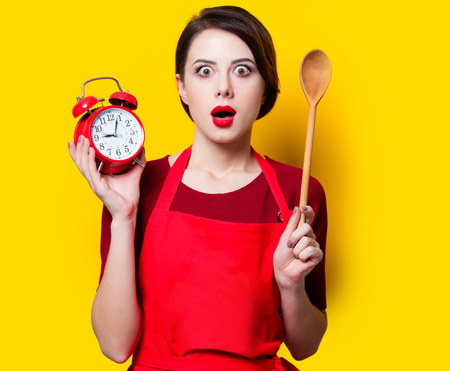 Portrait of young housewife with red alarm clock and spoon on yellow backgroundの写真素材