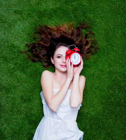 Portrait of a young redhead woman with vintage alarm clock lying down on green spring grass, above...