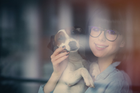 beautiful young woman standing near the window and hugging her dogの写真素材
