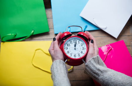 beautiful woman hands holding red alarm clock on the wonderful colorful shopping bag backgroundの写真素材