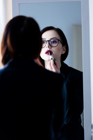 beautiful young woman standing in front of mirror, putting on lipstick and looking at her reflectionの写真素材