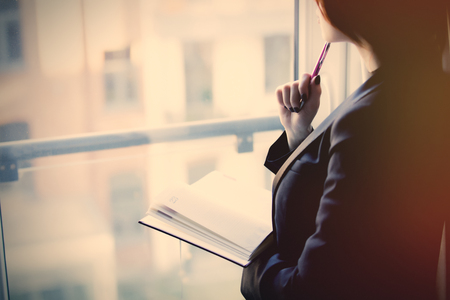 young beautiful woman holding a notebook and thinking near the windowの写真素材