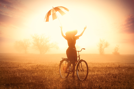 photo of beautiful young woman on bicycle with umbrella on the wonderful sunset backgroundの写真素材