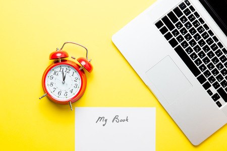photo of sheet of paper My Book, cool laptop and red alarm clock on the wonderful yellow studio backgroundの写真素材