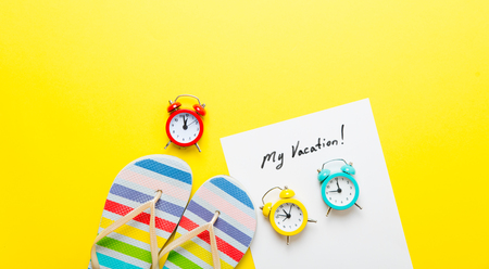 photo of sheet of paper My Vacation, colorful alarm clocks and colorful sandals on the wonderful yellow studio backgroundの写真素材