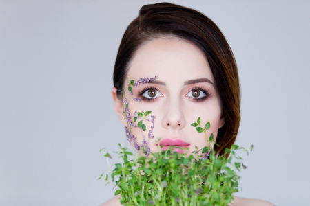 portrait of beautiful young woman with flowers on her face holding plant on the wonderful grey studio backgroundの写真素材